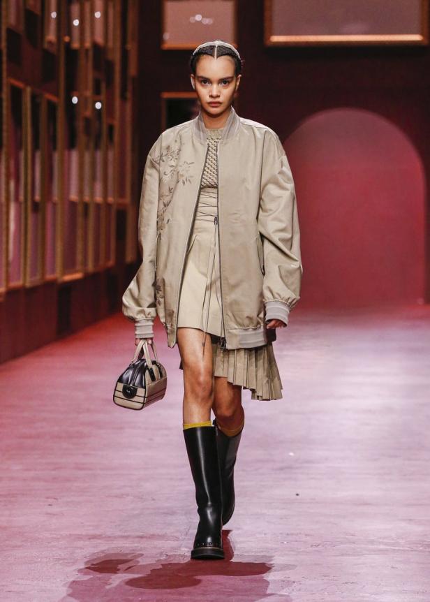 Eine Frau in einem beigen Ensemble mit Jacke, Rock, Stiefeln und Handtasche auf einem Laufsteg.