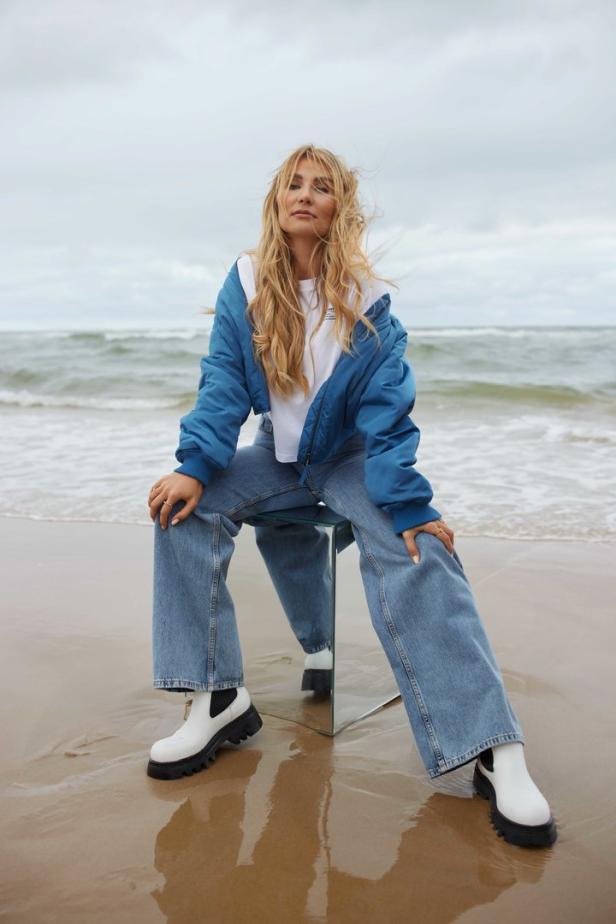 Eine Frau sitzt auf einem Stuhl am Strand, gekleidet in Jeans und einer blauen Jacke.