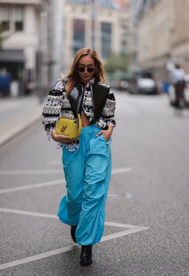 Eine Frau mit Sonnenbrille, hellblauer Hose und gelber Handtasche geht auf der Straße.