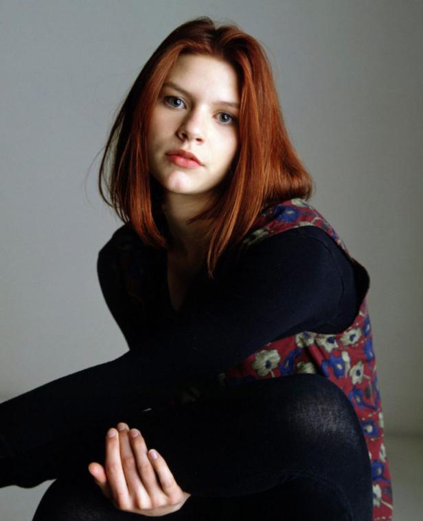 Claire Danes sitzt mit verschränkten Armen und rotem Haar vor einem grauen Hintergrund.