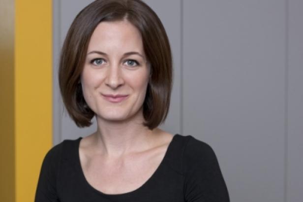Eine Frau mit braunen Haaren und einem schwarzen Oberteil lächelt in die Kamera.