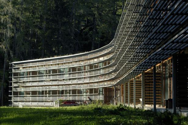 Ein modernes, geschwungenes Gebäude mit Holzfassade vor einem Wald.