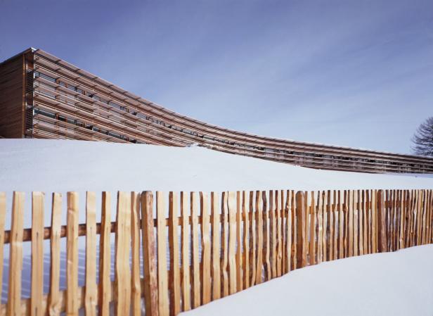Ein geschwungenes Holzgebäude hinter einem Holzzaun in einer verschneiten Landschaft.