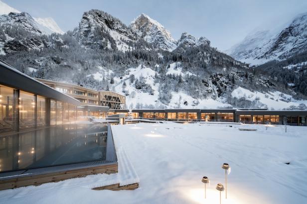 Ein modernes Hotel mit Außenpool liegt vor einer verschneiten Bergkulisse.