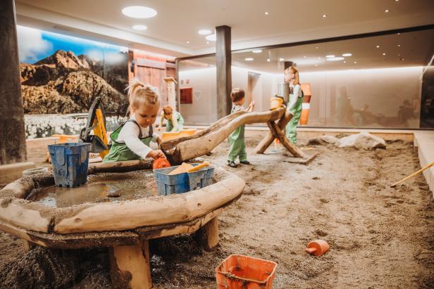 Mehrere Kinder spielen in einem Indoor-Sandkasten mit Wasser und Spielzeug.