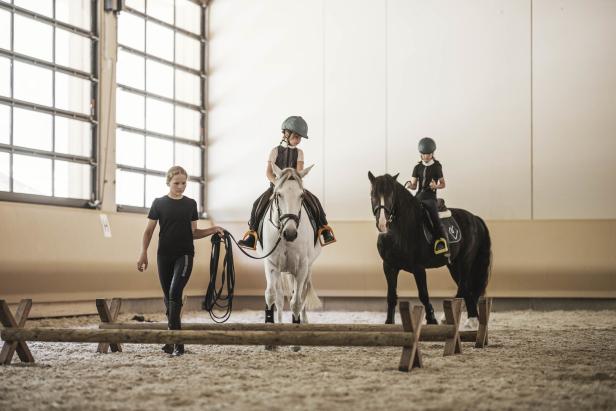 Zwei Kinder reiten auf Pferden in einer Reithalle, während ein Mädchen daneben steht.