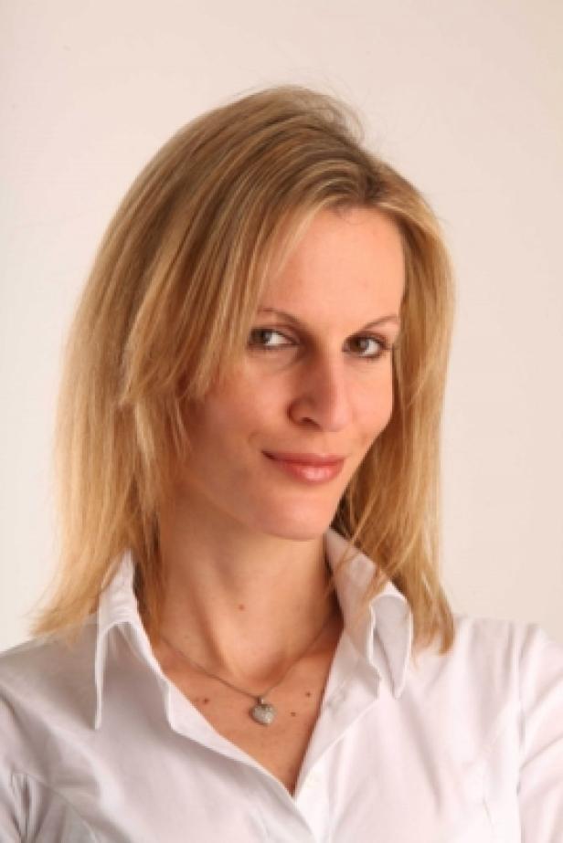 Eine blonde Frau mit weißem Hemd und einer Halskette lächelt.