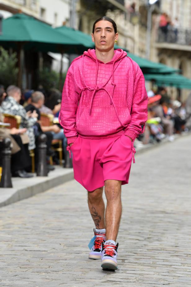 Ein Mann in einem pinkfarbenen Hoodie und Shorts läuft auf einem Laufsteg.
