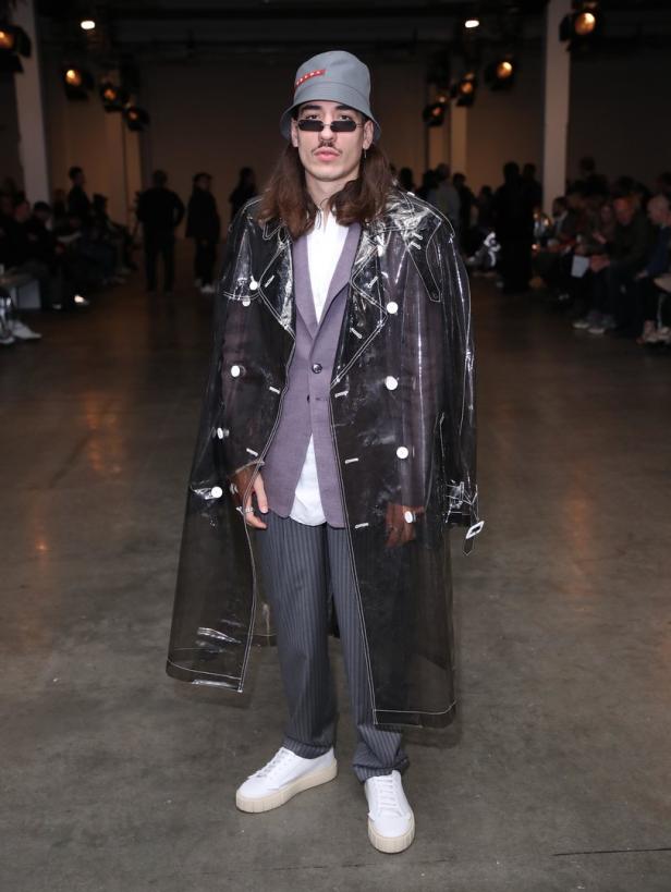 Ein Mann mit Hut und Sonnenbrille trägt einen transparenten Trenchcoat.