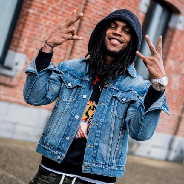 Ein lächelnder Mann mit Dreadlocks, Kapuzenpullover und Jeansjacke zeigt Handzeichen vor einer Backsteinmauer.