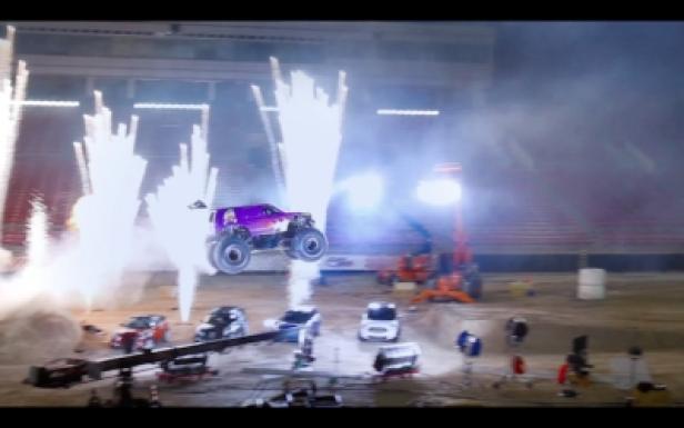 Ein lila Monster Truck springt über mehrere Autos, begleitet von Feuerwerk.