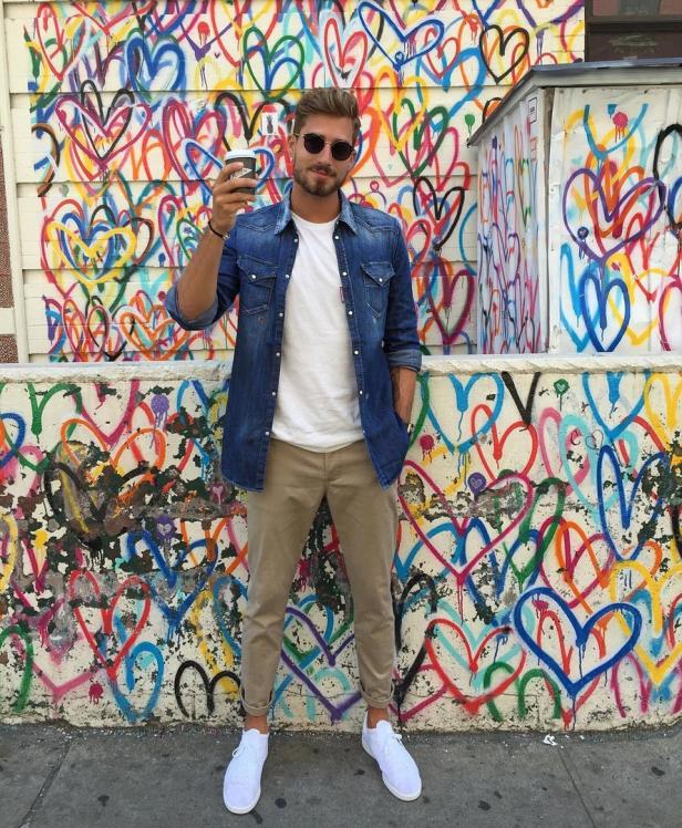 Ein Mann mit Sonnenbrille posiert mit einem Kaffee vor einer Wand mit Graffiti-Herzen.