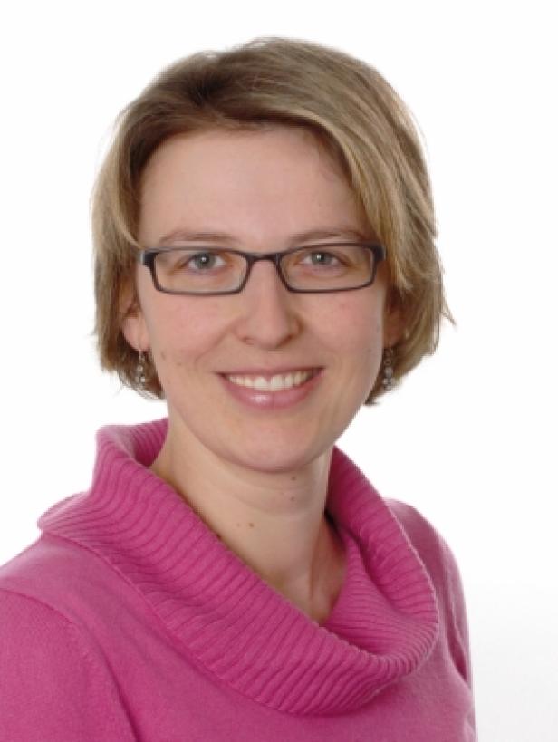 Eine lächelnde Frau mit Brille und pinkfarbenem Pullover vor einem weißen Hintergrund.