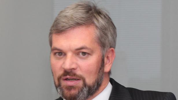 Ein Mann mit grau meliertem Haar und Bart in einem dunklen Anzug.