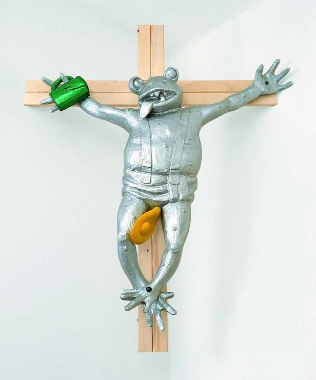 Eine silberne Frosch-Figur in Latzhose hängt an einem Holzkreuz.