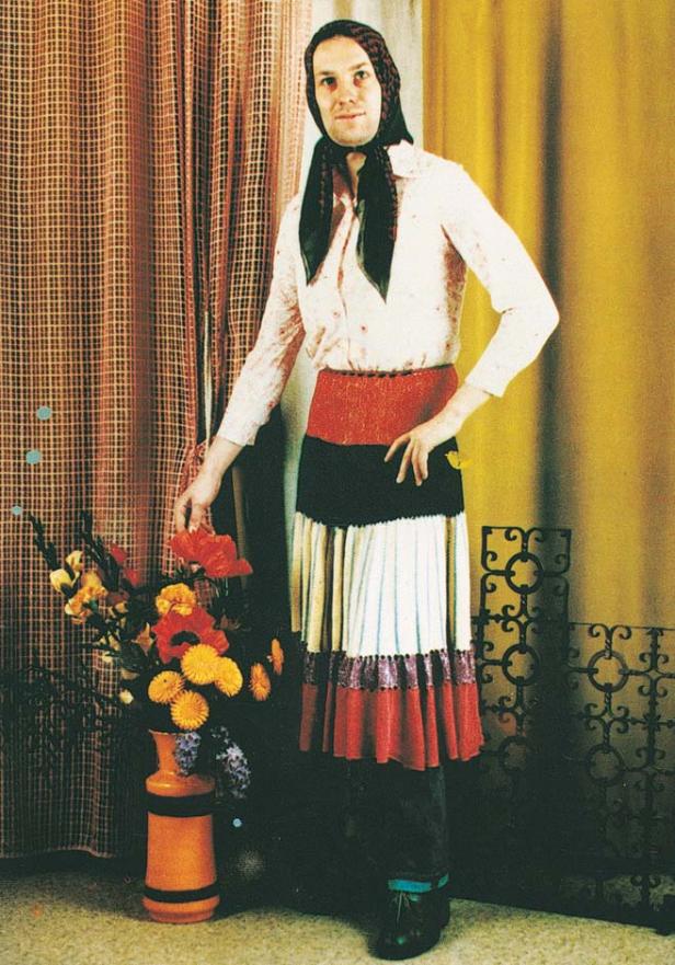 Ein Mann mit Kopftuch, Bluse und Rock posiert neben einem Blumenarrangement.