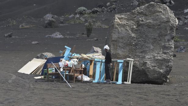 Eine Person steht neben einem Haufen aus Holz und anderen Materialien auf schwarzem Sand.