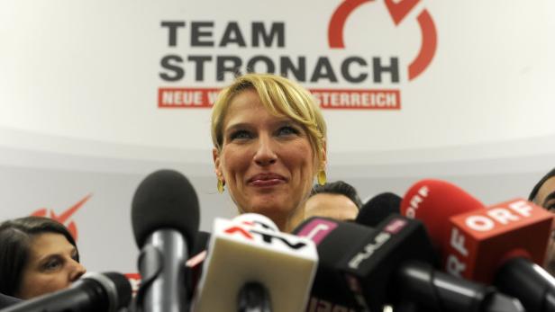 Eine Frau steht vor Mikrofonen mit dem Logo von Team Stronach im Hintergrund.