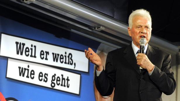Ein älterer Mann spricht in ein Mikrofon vor einem Schild mit der Aufschrift „Weil er weiß, wie es geht“.