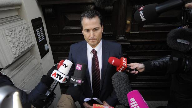 Ein Mann in Anzug steht vor einer Tür und wird von Reportern mit Mikrofonen umringt.