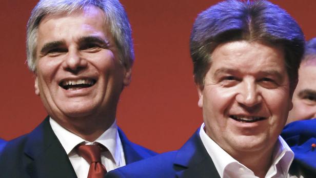 Zwei lächelnde Männer in Anzügen stehen vor einem roten Hintergrund.