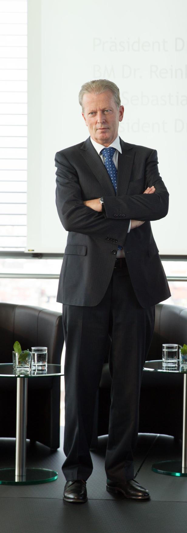 Ein Mann im Anzug steht mit verschränkten Armen vor einem Fenster.