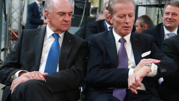 Zwei Männer in Anzügen sitzen nebeneinander bei einer Veranstaltung.