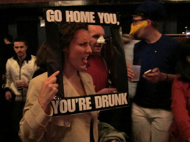 Eine Frau mit einem Schild um den Hals, auf dem „Go Home You, You're Drunk“ steht, gestikuliert in einem Raum voller Menschen.