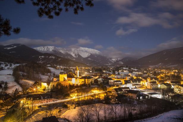 Blick auf eine Stadt mit beleuchteten Gebäuden und schneebedeckten Bergen im Hintergrund bei Nacht.