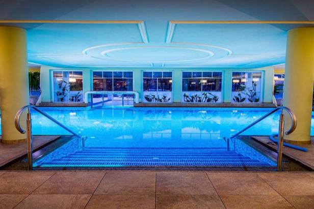 Ein Schwimmbecken in einem Innenraum mit blauer Beleuchtung und großen Fenstern.
