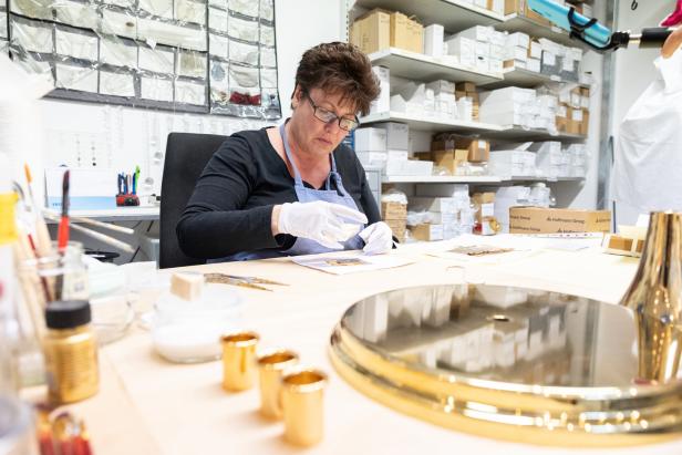 Eine Frau mit Handschuhen arbeitet an einem Tisch mit kleinen goldenen Gegenständen.