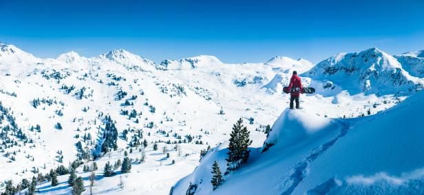 Ein Snowboarder steht auf einem Gipfel mit Blick auf eine verschneite Berglandschaft.