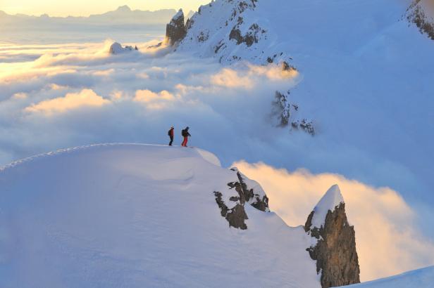 Zwei Skifahrer stehen auf einem schneebedeckten Gipfel mit Blick auf eine Wolkendecke.