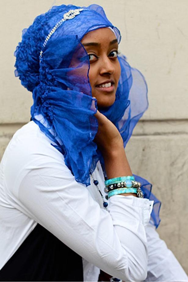 Eine junge Frau mit blauem Hijab und Armbändern lächelt in die Kamera.