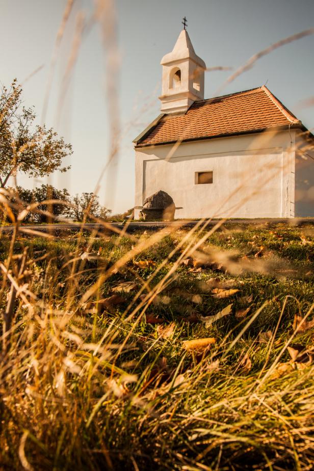 Eine kleine, weiße Kapelle mit einem roten Ziegeldach auf einem Hügel im Herbst.