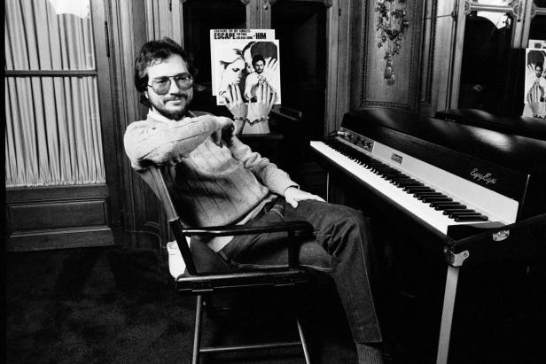 Rupert Holmes sitzt neben einem Rhodes-Piano und hält sein Album „Escape“ hoch.