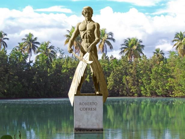 Eine Statue von Roberto Cofresí steht in einem Gewässer, umgeben von Palmen und Bäumen.