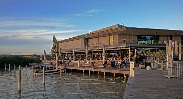 Ein Restaurant am Wasser mit Gästen auf einer Terrasse bei Sonnenuntergang.