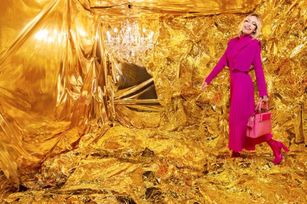 Eine Frau in einem pinkfarbenen Outfit posiert vor einem goldenen Hintergrund mit einem Kronleuchter.