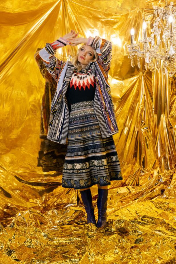 Eine Frau in einem bunten Outfit posiert vor einem goldenen Hintergrund mit einem Kristalllüster.