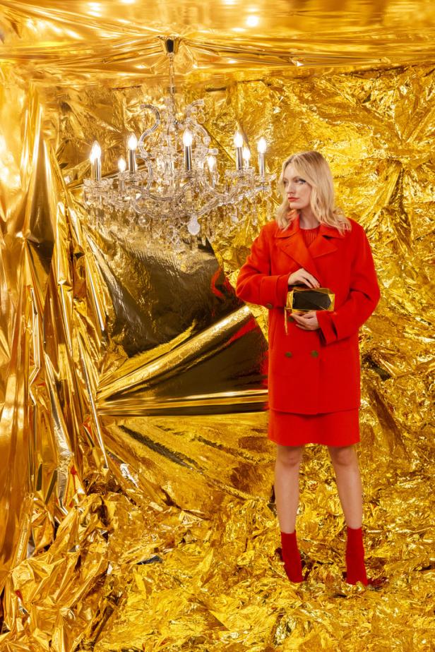 Eine Frau in einem roten Ensemble posiert vor einem goldenen Hintergrund mit einem Kronleuchter.