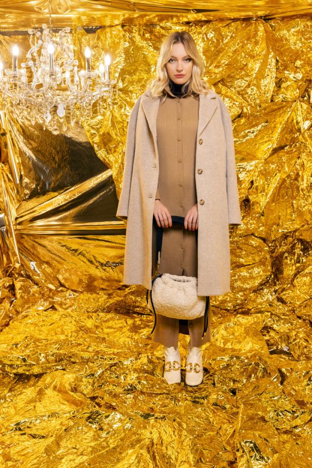 Eine Frau in einem beigen Mantel und Kleid vor einem goldenen Hintergrund mit einem Kronleuchter.