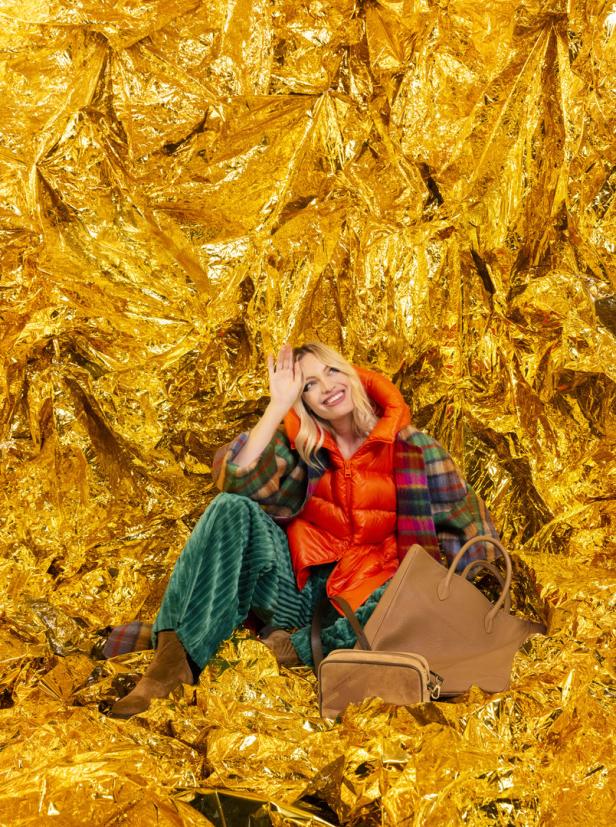Eine Frau sitzt vor einem goldenen Hintergrund und trägt eine orangefarbene Weste und grüne Hosen.