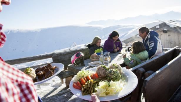 Ein Kellner serviert Essen an einem Tisch mit einer Familie vor einer verschneiten Berglandschaft.