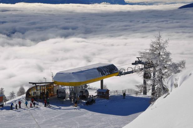 Eine Gondelbahnstation auf einem Berg mit Skifahrern und einer Wolkendecke im Hintergrund.