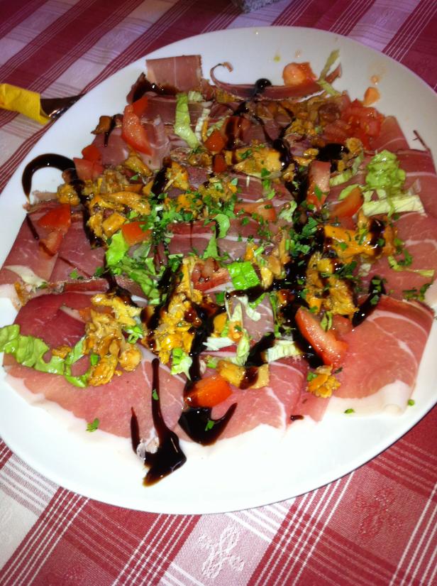 Ein Teller mit Schinken, Pfifferlingen, Tomaten und Balsamico-Glasur auf einem rot-weiß karierten Tisch.