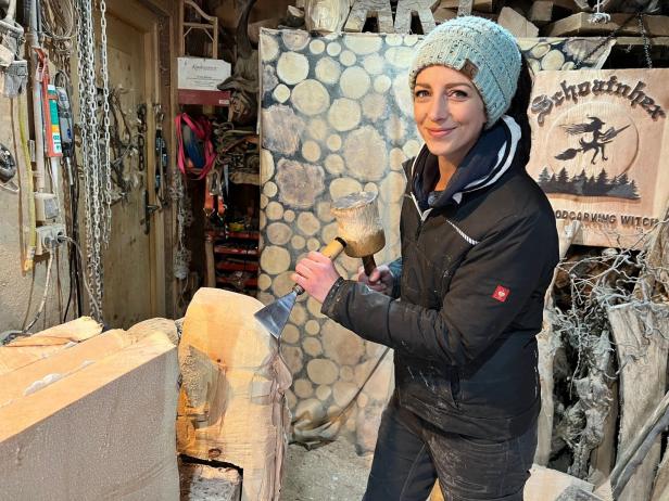 Eine Frau meißelt mit einem Klopfholz eine Holzskulptur in ihrer Werkstatt.