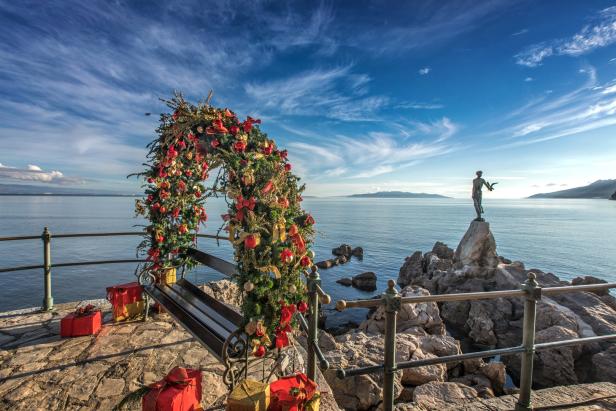 Ein mit Weihnachtsschmuck dekorierter Bogen und Geschenke vor einer Küstenlandschaft mit einer Statue.
