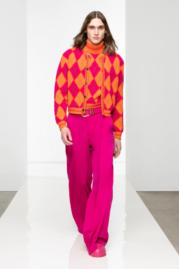 Ein schlanker Mann mit langen Haaren trägt eine orange-pinkfarbene Argyle-Strickjacke und eine pinkfarbene Hose.