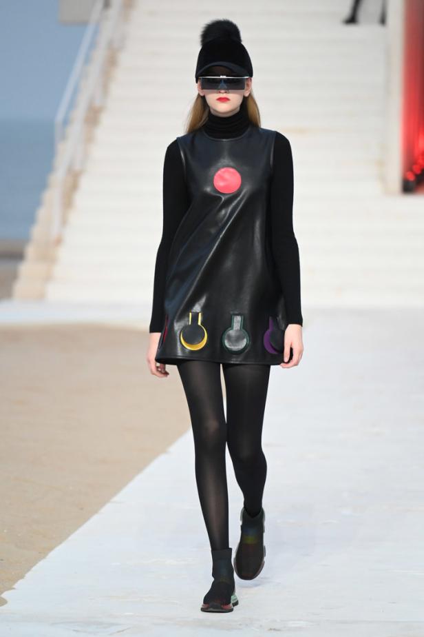 Eine Frau mit futuristischer Brille und Mütze präsentiert ein schwarzes Kleid mit bunten Applikationen.